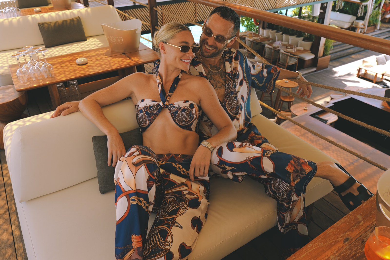 Couple in matching Hustle printed satin co-ord sets lounging on a beach club daybed — his & hers looks for Ibiza days